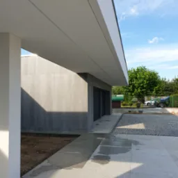 Nowoczesny, minimalistyczny dom z szarą elewacją i białym zadaszeniem, widok z zewnątrz na świeżo ułożony chodnik z kostki brukowej i płyt kamiennych, błękitne niebo w tle.