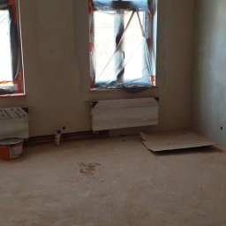 REMONTY DAMIAN DUSZCZAK - Pokój w trakcie prac tynkarskich: ściany pokryte świeżym tynkiem, okna zabezpieczone folią, grzejniki osłonięte, widoczne narzędzia i materiały budowlane w rogu pomieszczenia.