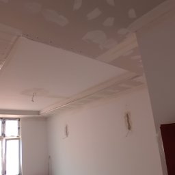 REMONTY DAMIAN DUSZCZAK - Montaż podwieszanego sufitu z płyt gipsowo-kartonowych, widoczne profile i wstępnie zaszpachlowane łączenia, surowy stan wnętrza z oknem w tle.