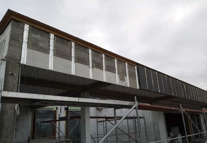 Budynek w trakcie ocieplania elewacji, widoczne płyty izolacyjne i rusztowania, szara betonowa konstrukcja w tle pochmurnego nieba.