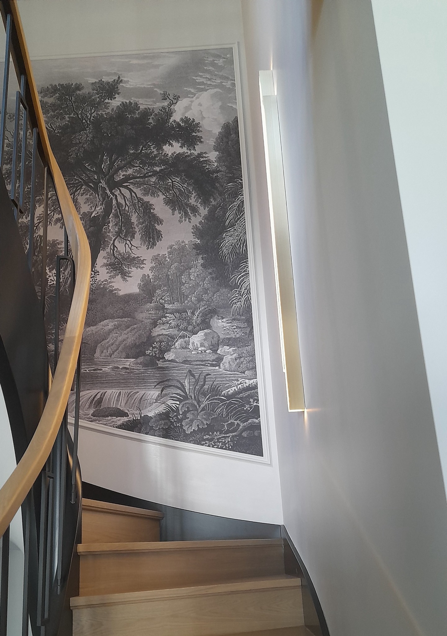 Klatka schodowa z drewnianymi schodami, czarnymi metalowymi balustradami z drewnianym poręczą oraz dużym czarno-białym obrazem krajobrazu na ścianie i nowoczesnym oświetleniem ściennym.