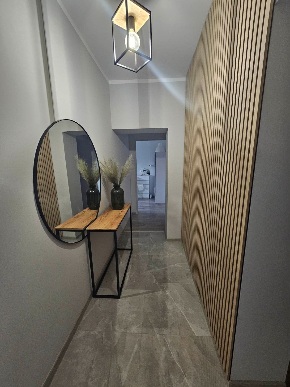 Nowoczesna aranżacja przedpokoju w Brodnicy: lamelowa ściana w naturalnym odcieniu drewna, podłoga z szarych płytek imitujących marmur, okrągłe lustro nad drewnianą konsolą z czarnymi nogami...