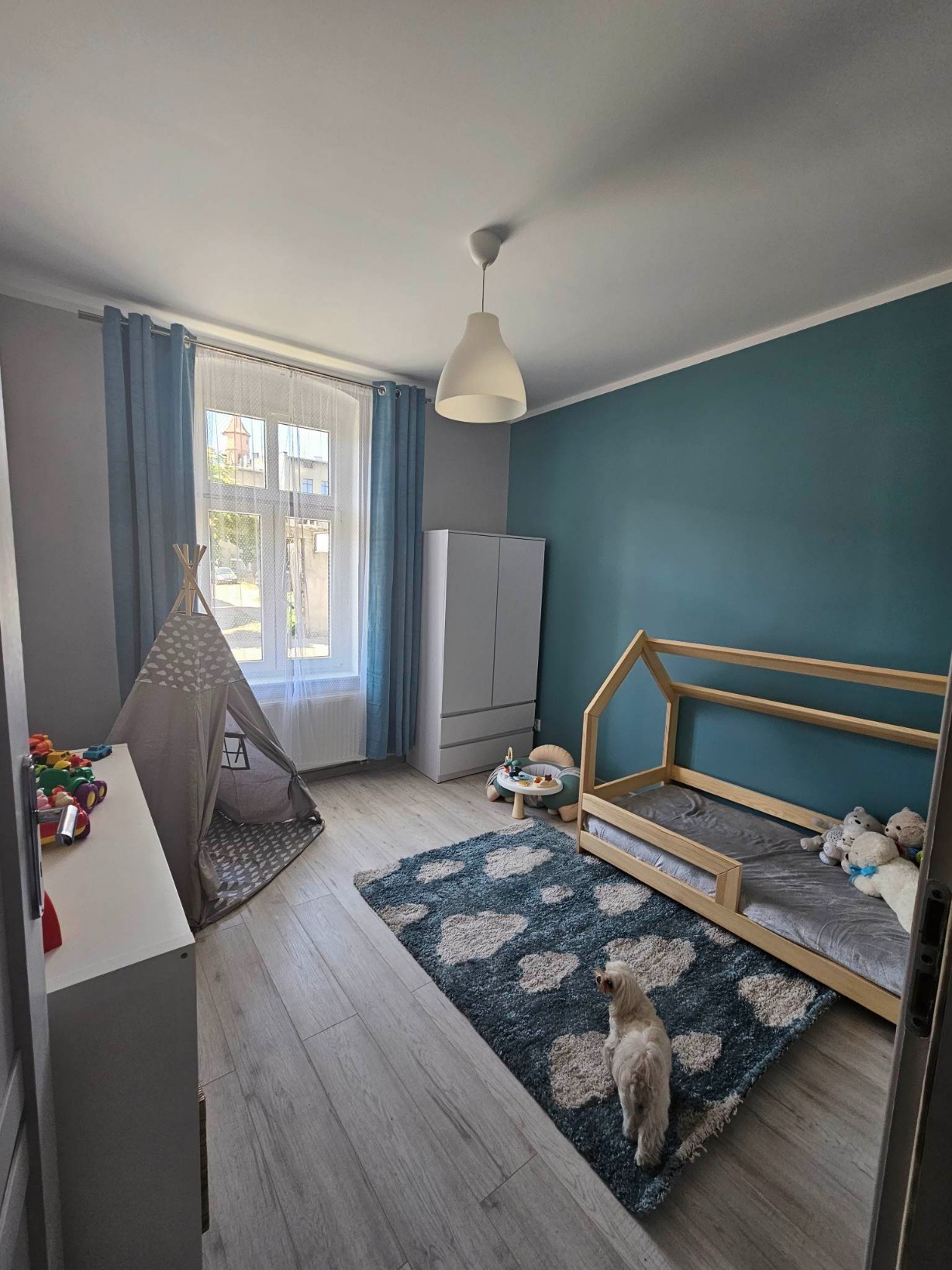 Umeblowany pokój dziecięcy z łóżkiem w kształcie domku, tipi, białymi meblami, błękitnymi ścianami i dywanem w chmurki, oświetlony naturalnym światłem z okna, z małym psem na dywanie.