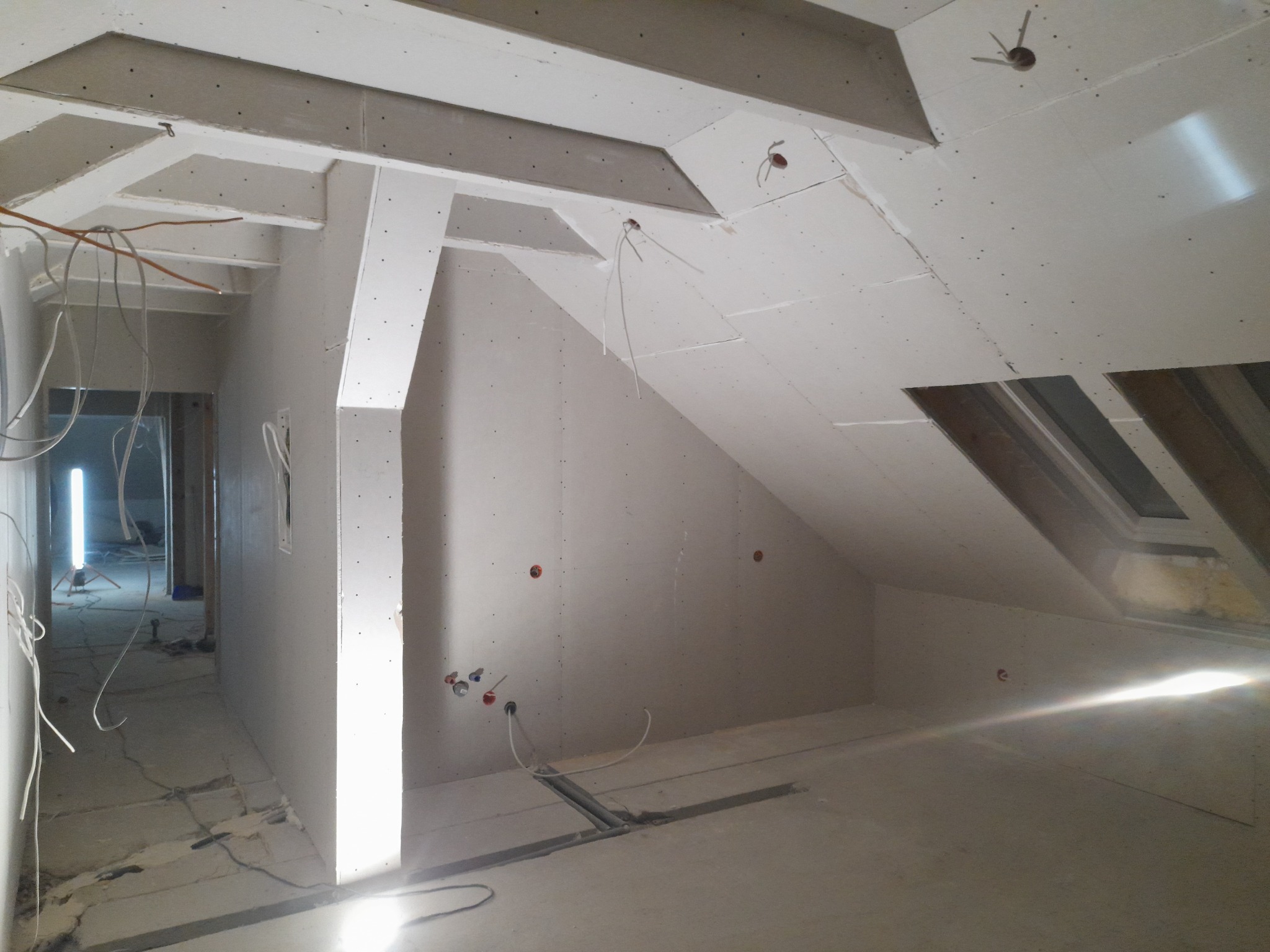 Poddasze w trakcie wykańczania płytami kartonowo-gipsowymi, widoczne stelaże, instalacje elektryczne i okno dachowe, stan surowy przed malowaniem.