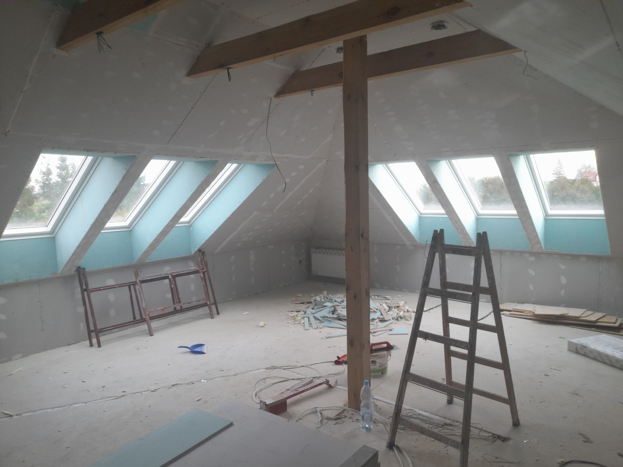 Poddasze w trakcie wykańczania z widocznymi oknami dachowymi, konstrukcją drewnianą, częściowo położonymi płytami kartonowo-gipsowymi oraz rozłożonymi narzędziami i materiałami budowlanymi.