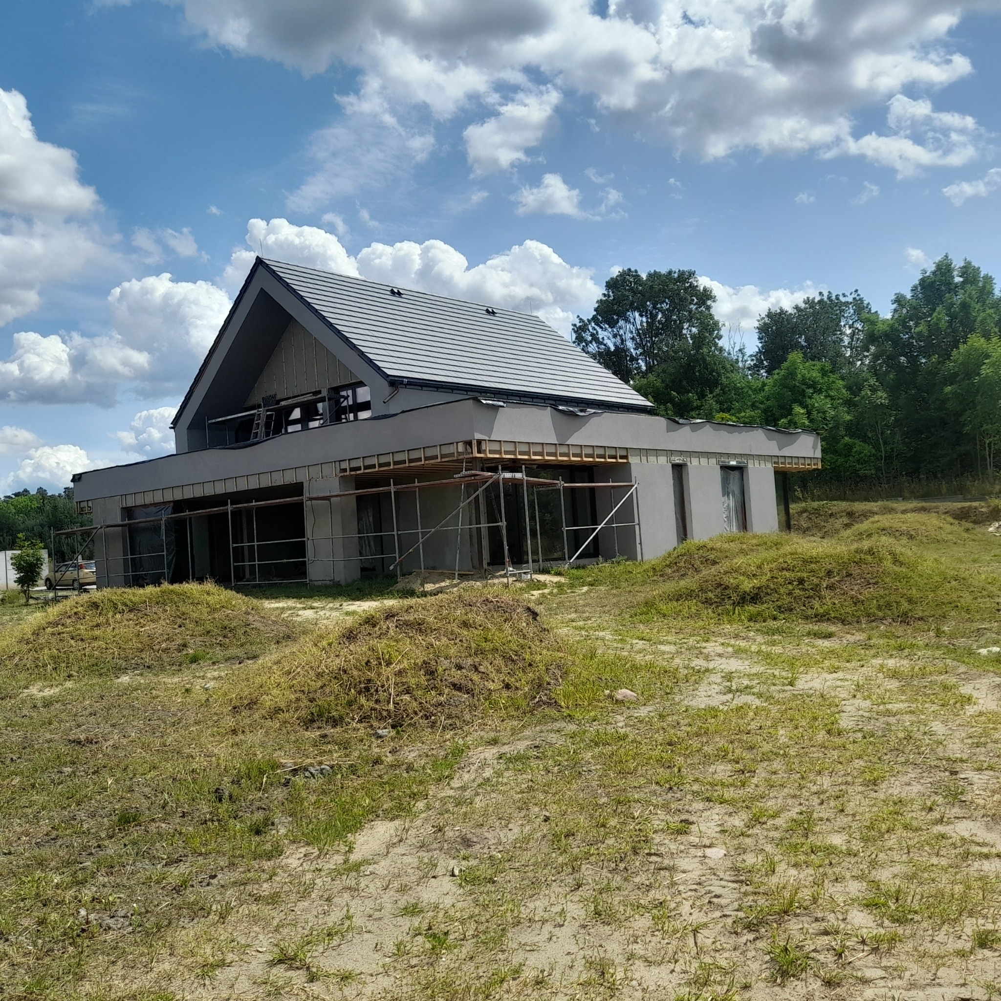 Nowoczesny dom jednorodzinny w trakcie wykończenia elewacji, widoczne rusztowania i szara kolorystyka budynku na tle błękitnego nieba z chmurami.