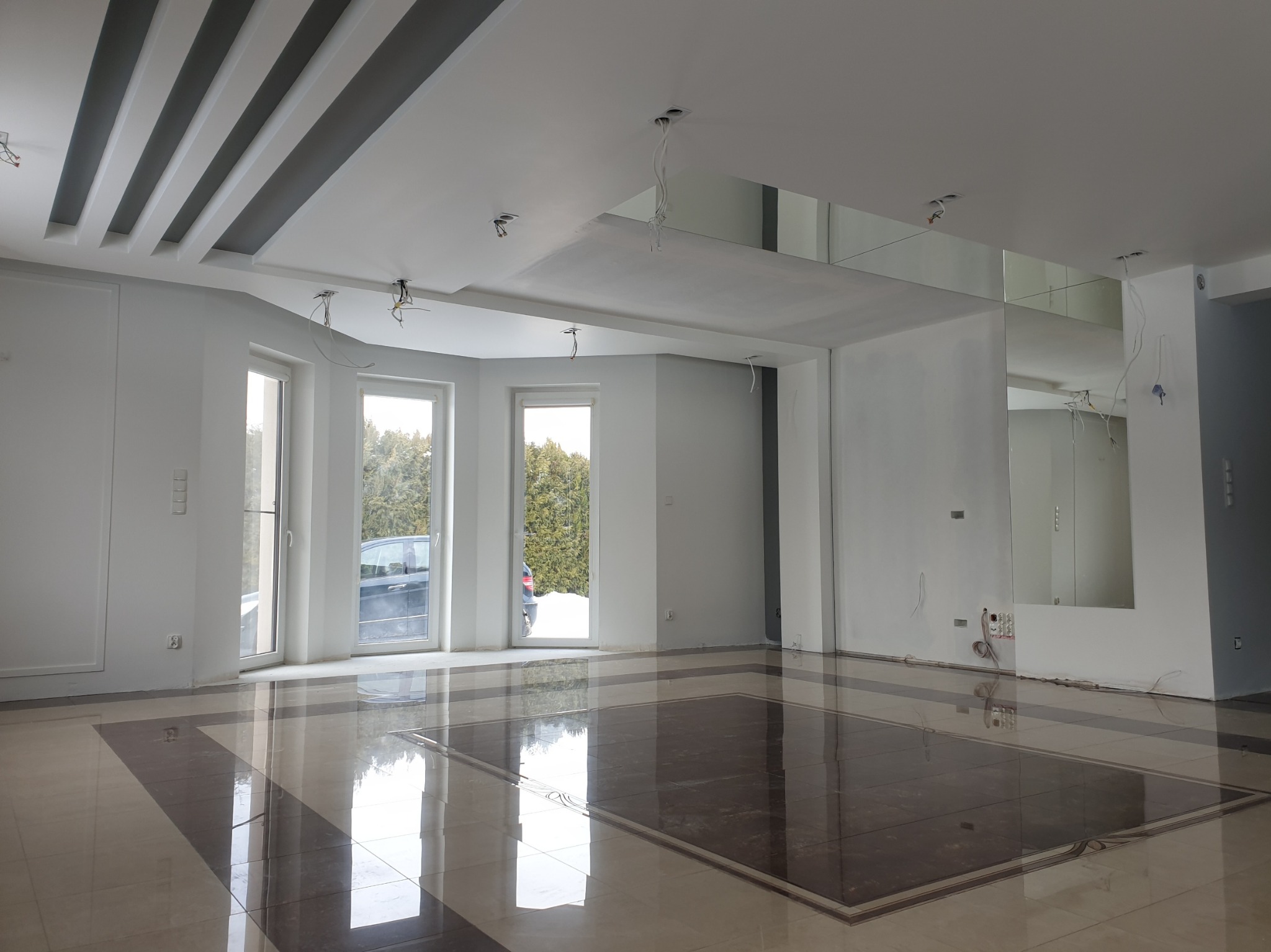 Przestronne, jasne wnętrze w trakcie wykańczania: białe ściany, błyszcząca podłoga z płytek w geometryczny wzór, sufit z dekoracyjnymi, szarymi belkami i lustrami, widoczne kable wystające z sufitu...