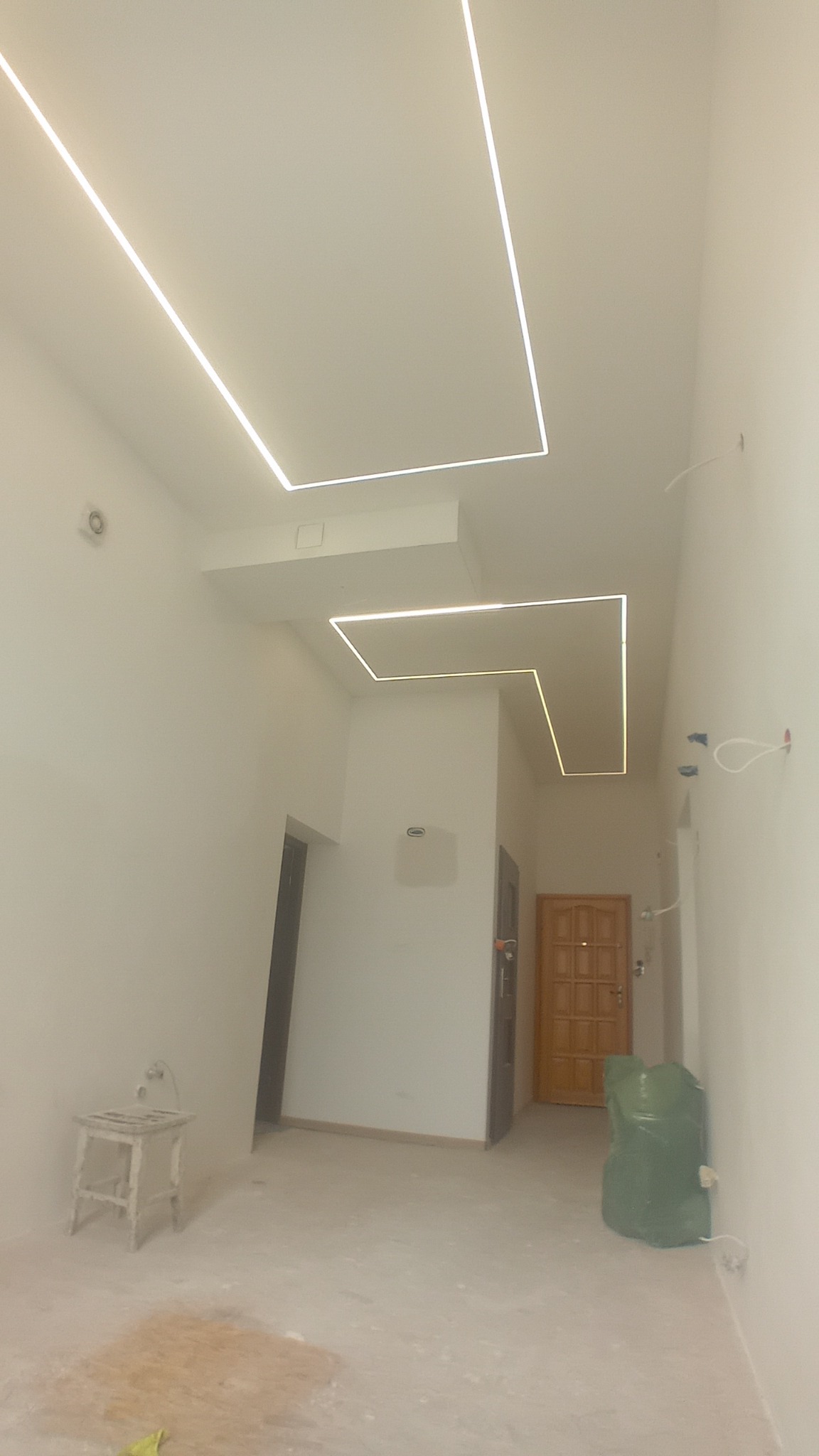 Wnętrze w trakcie prac wykończeniowych: surowe ściany, podłoga z resztkami materiałów, nowoczesne oświetlenie LED w geometrycznych kształtach na suficie, widoczne drzwi wejściowe i wewnętrzne.