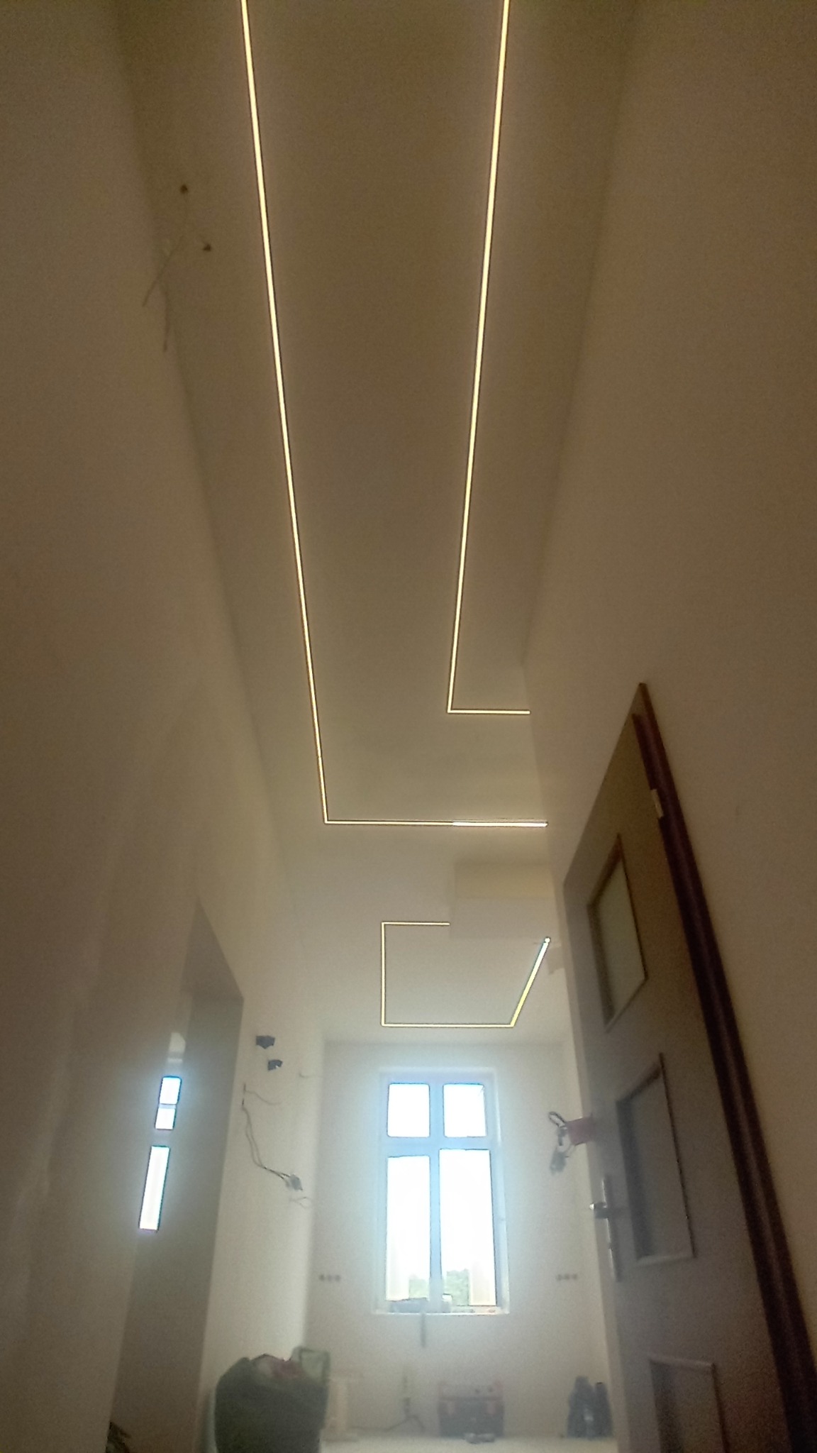 Korytarz w trakcie prac wykończeniowych z zamontowanym, nowoczesnym oświetleniem LED wpuszczonym w sufit, widoczne niedociągnięcia i przewody na ścianach.