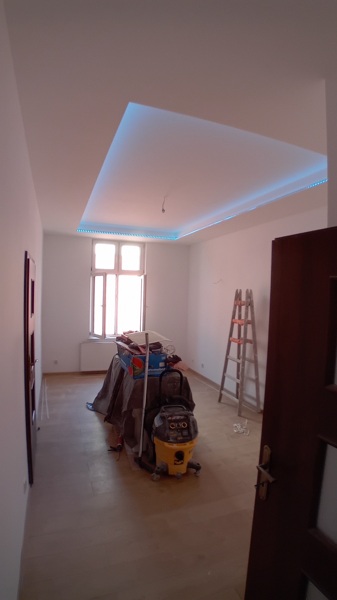Pokój w trakcie odświeżania: widoczny nowy sufit z oświetleniem LED, drabina, odkurzacz przemysłowy i zabezpieczone folią meble.