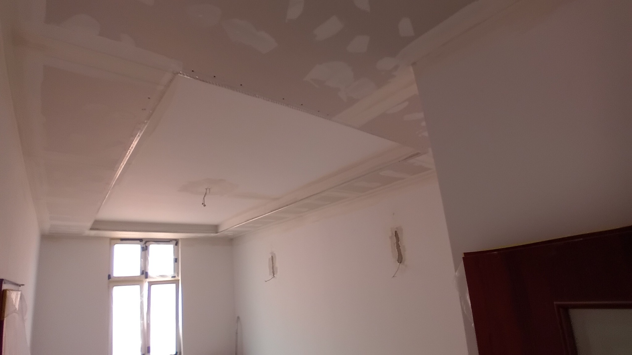 Montaż podwieszanego sufitu z płyt gipsowo-kartonowych, widoczne profile i wstępnie zaszpachlowane łączenia, surowy stan wnętrza z oknem w tle.