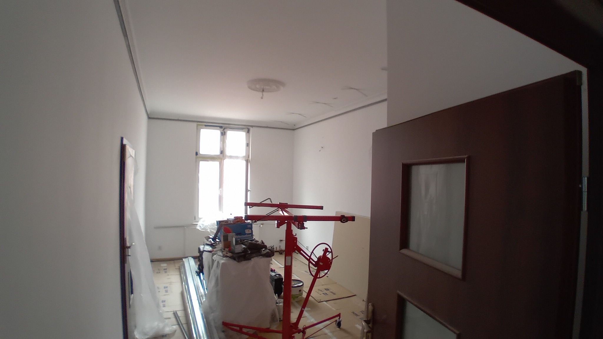 Pokój w trakcie prac wykończeniowych: czerwony podnośnik do płyt gipsowo-kartonowych, zabezpieczona folią podłoga, świeżo pomalowane ściany i sufit z widocznym ornamentem, okno z białymi ramami...