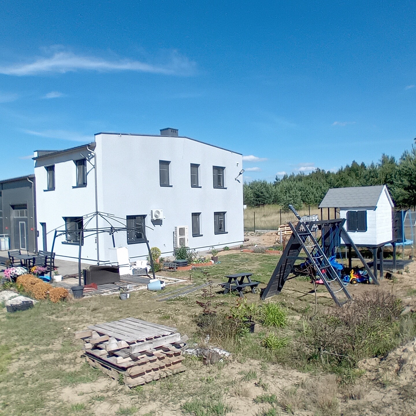 Dwupiętrowy dom z szarą elewacją i ciemnymi obramowaniami okien, widok na plac zabaw z drewnianą konstrukcją i małym domkiem, palety na trawie, w tle las i błękitne niebo.
