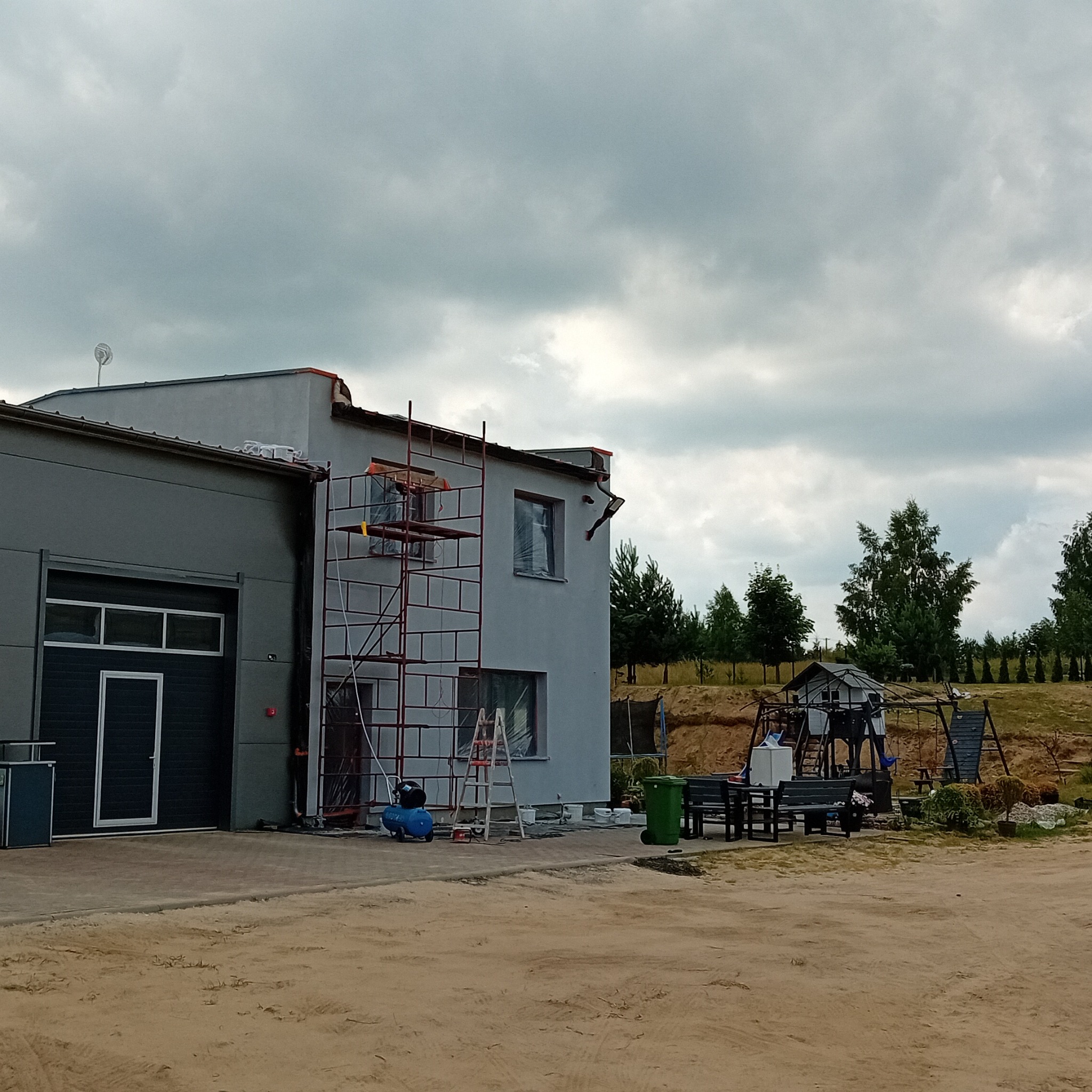 Dwukondygnacyjny budynek z szarą elewacją w trakcie prac wykończeniowych, z widocznym rusztowaniem, sprężarką i drabina opartą o ścianę, z placem zabaw w tle.