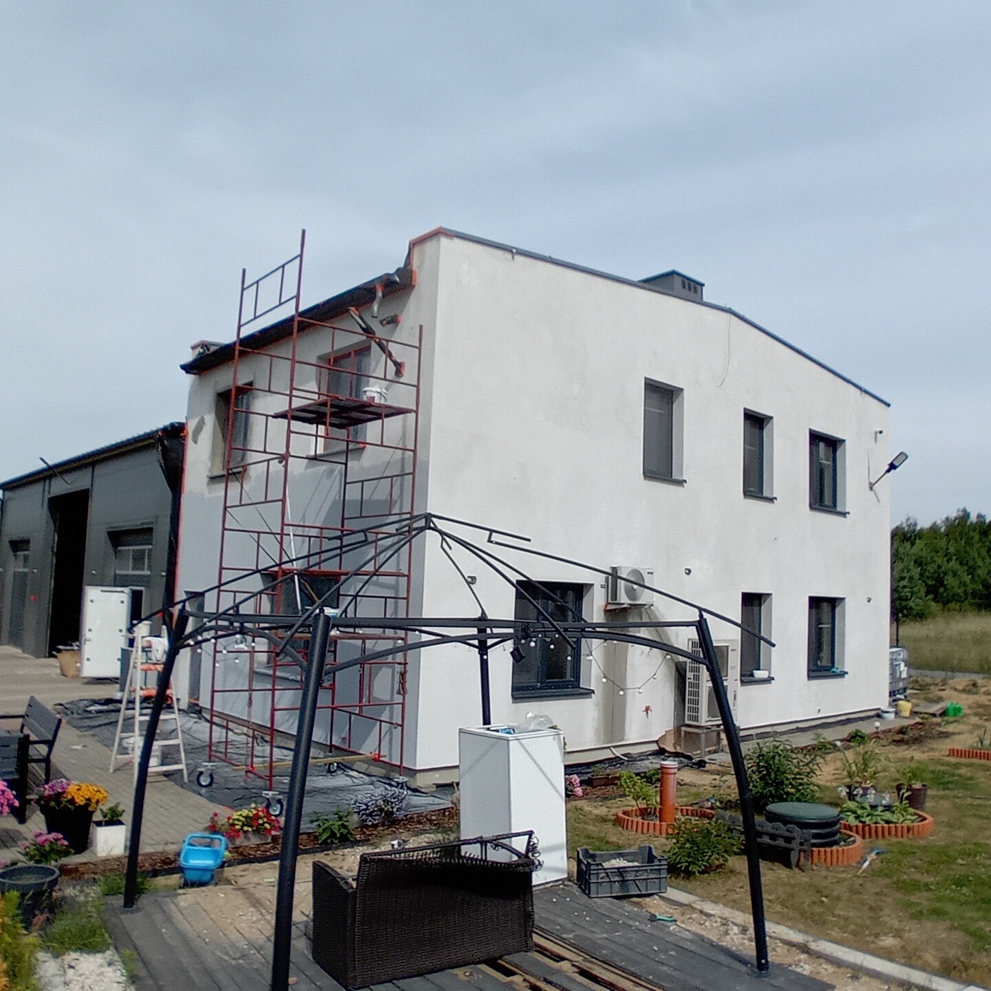 Biały dom z szarymi oknami, w trakcie prac na elewacji, widoczne rusztowania, klimatyzatory na ścianie, częściowo zadaszony taras w budowie, kwiaty w donicach i zabawki na zewnątrz.