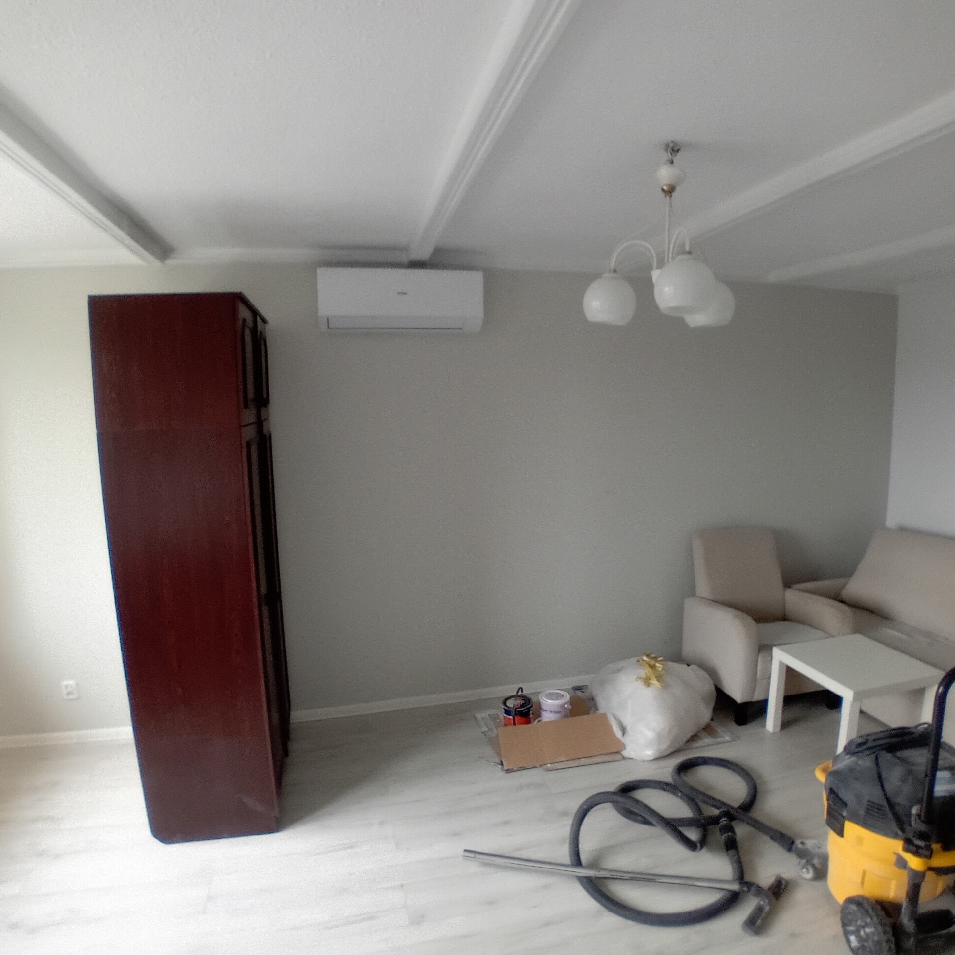 Pokój w trakcie remontu: pomalowane ściany na szaro, widoczny klimatyzator, meble pod folią ochronną, narzędzia i odkurzacz przemysłowy na podłodze.