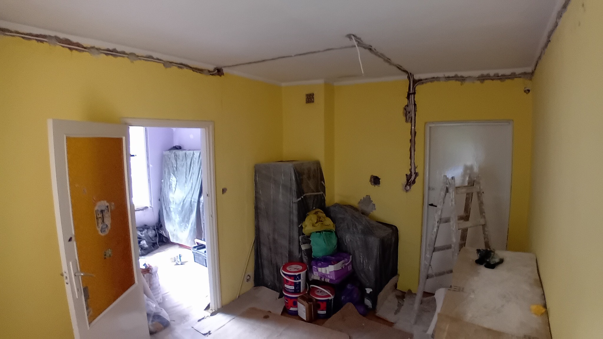 Pokój z żółtymi ścianami w trakcie prac przygotowawczych: widoczne kable elektryczne, ślady po kuciu, zakryte meble, drabina i farby.