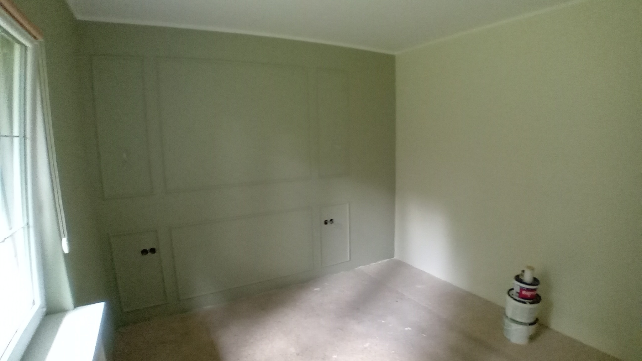 Pokój po malowaniu ścian na jasny odcień zieleni z dekoracyjnymi panelami ściennymi i ukrytymi drzwiczkami rewizyjnymi; widoczne puszki z farbą w rogu.