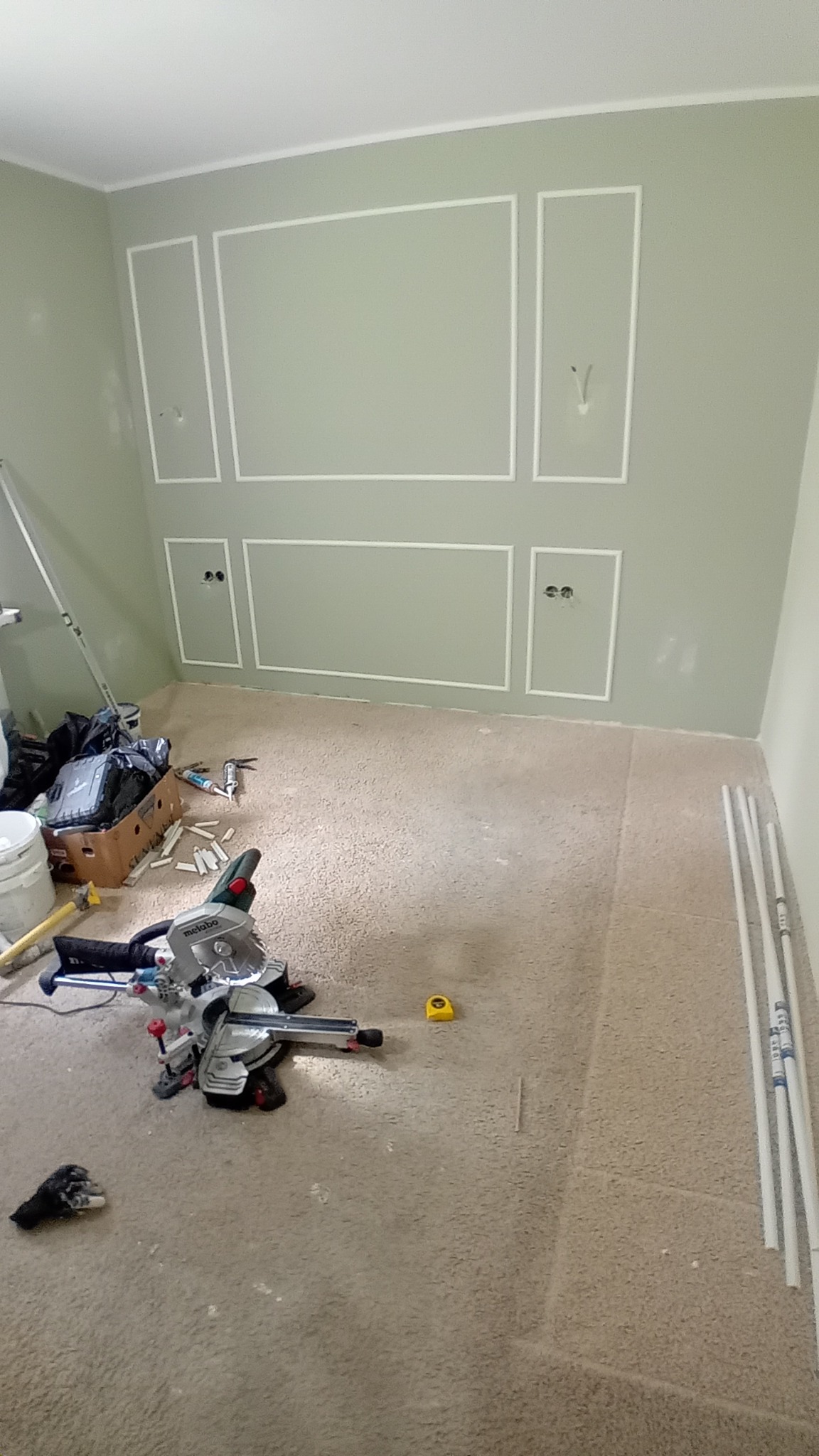 Montaż ozdobnych listew ściennych w pokoju z zielonymi ścianami, widoczna piła ukośnica, listwy przypodłogowe i narzędzia.