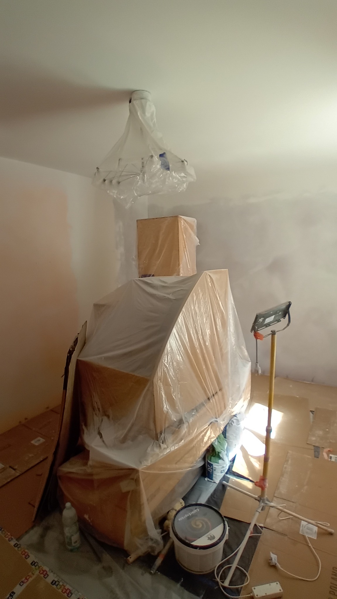 Przygotowania do malowania pokoju: meble zabezpieczone folią, lampa owinięta, widoczna puszka z farbą i lampa robocza.