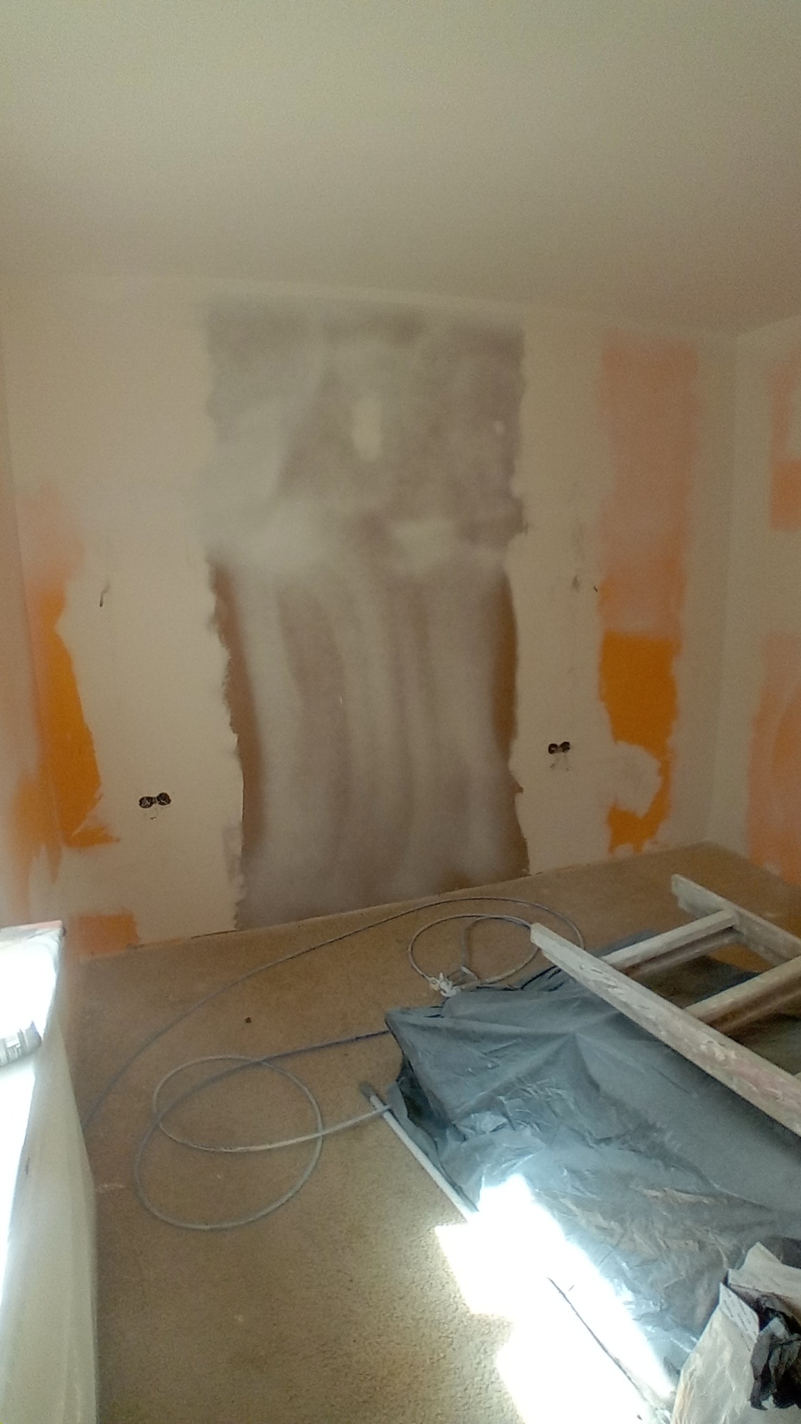 Pokój w trakcie przygotowania do malowania: częściowo zagruntowana ściana, zabezpieczone gniazdka, przewody na podłodze, drabina i folia ochronna.