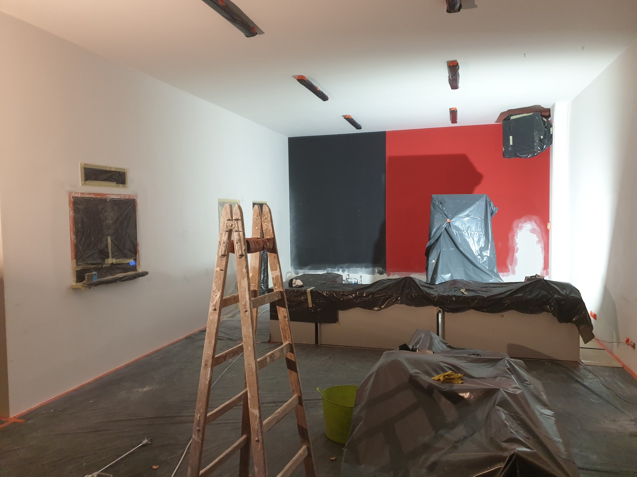 Pokój w trakcie malowania: ściany częściowo pomalowane na biało, czarno i czerwono, zabezpieczone okna, podłoga i meble przykryte folią, widoczna drewniana drabina i wiadro.