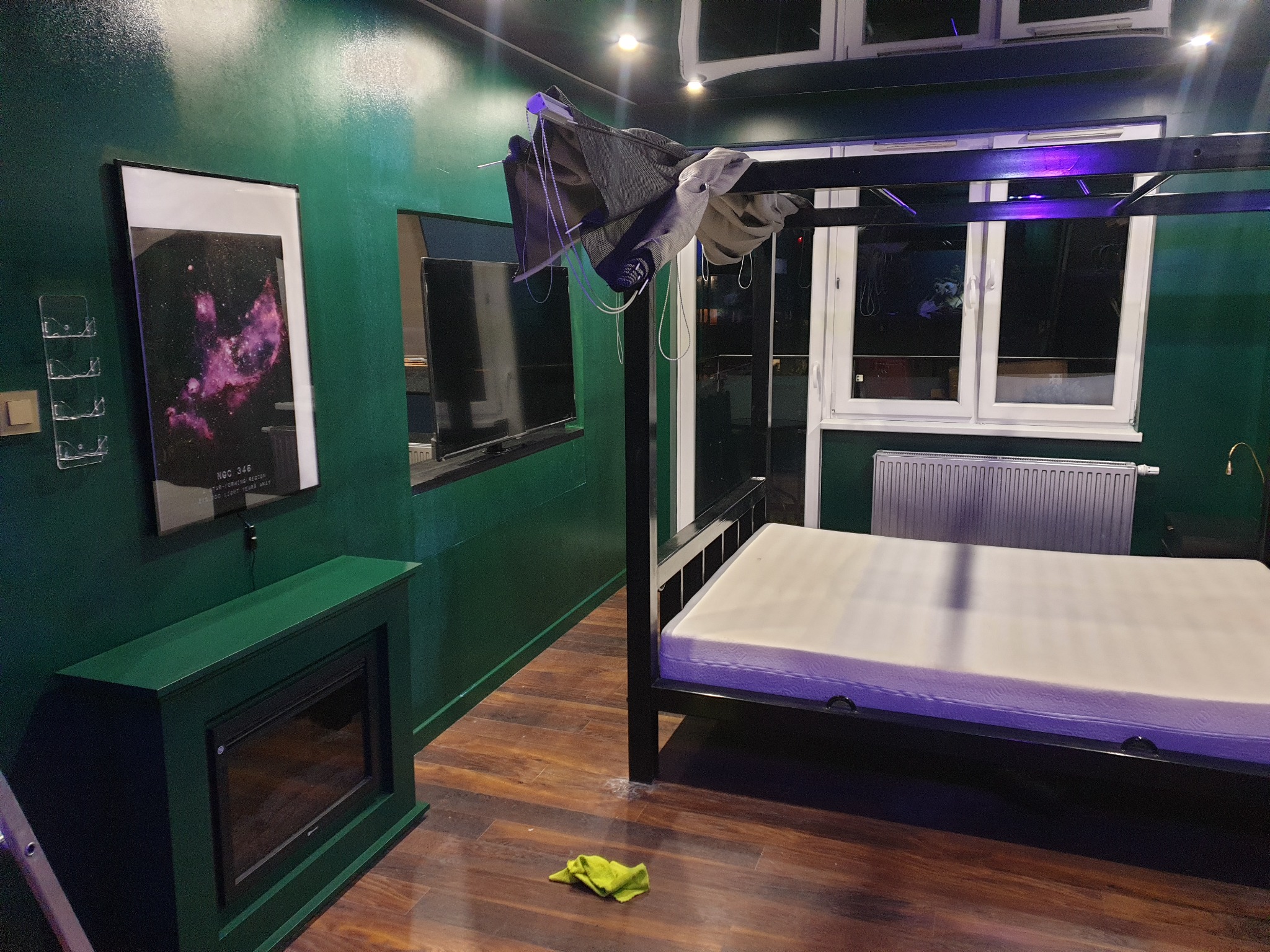 Pokój z ciemnozielonymi ścianami, czarnym łóżkiem z baldachimem bez pościeli, elektrycznym kominkiem i plakatem z galaktyką na ścianie.