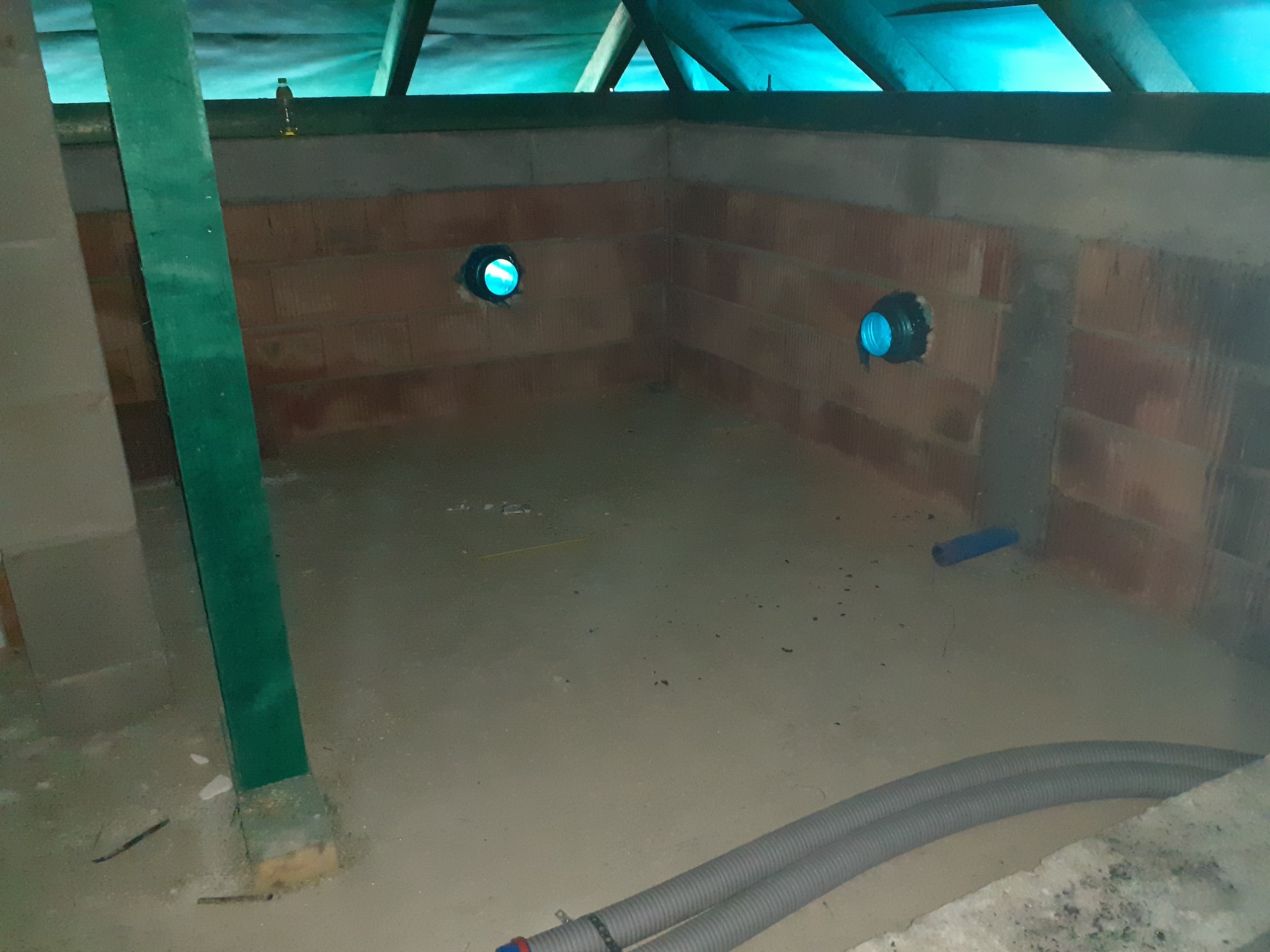 Surowa przestrzeń poddasza z dwoma otworami wentylacyjnymi w ścianie z czerwonej cegły, widoczne szare rury wentylacyjne na podłodze i zielona drewniana belka konstrukcyjna.