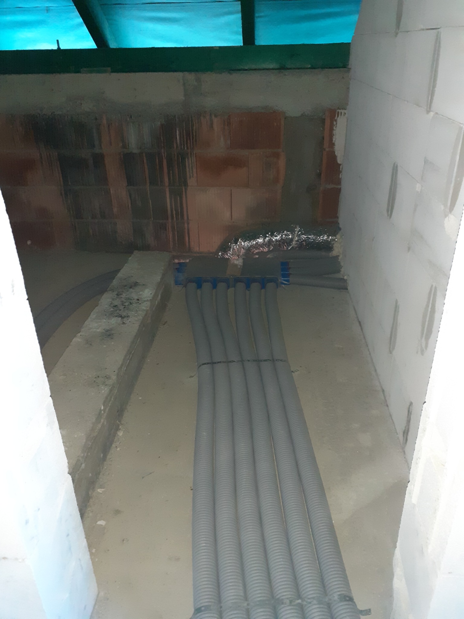 Szereg szarych, karbowanych rur wentylacyjnych z niebieskimi złączkami, ułożonych równolegle w surowym pomieszczeniu w trakcie instalacji; widoczne ściany z betonu komórkowego i cegieł, oraz folia...