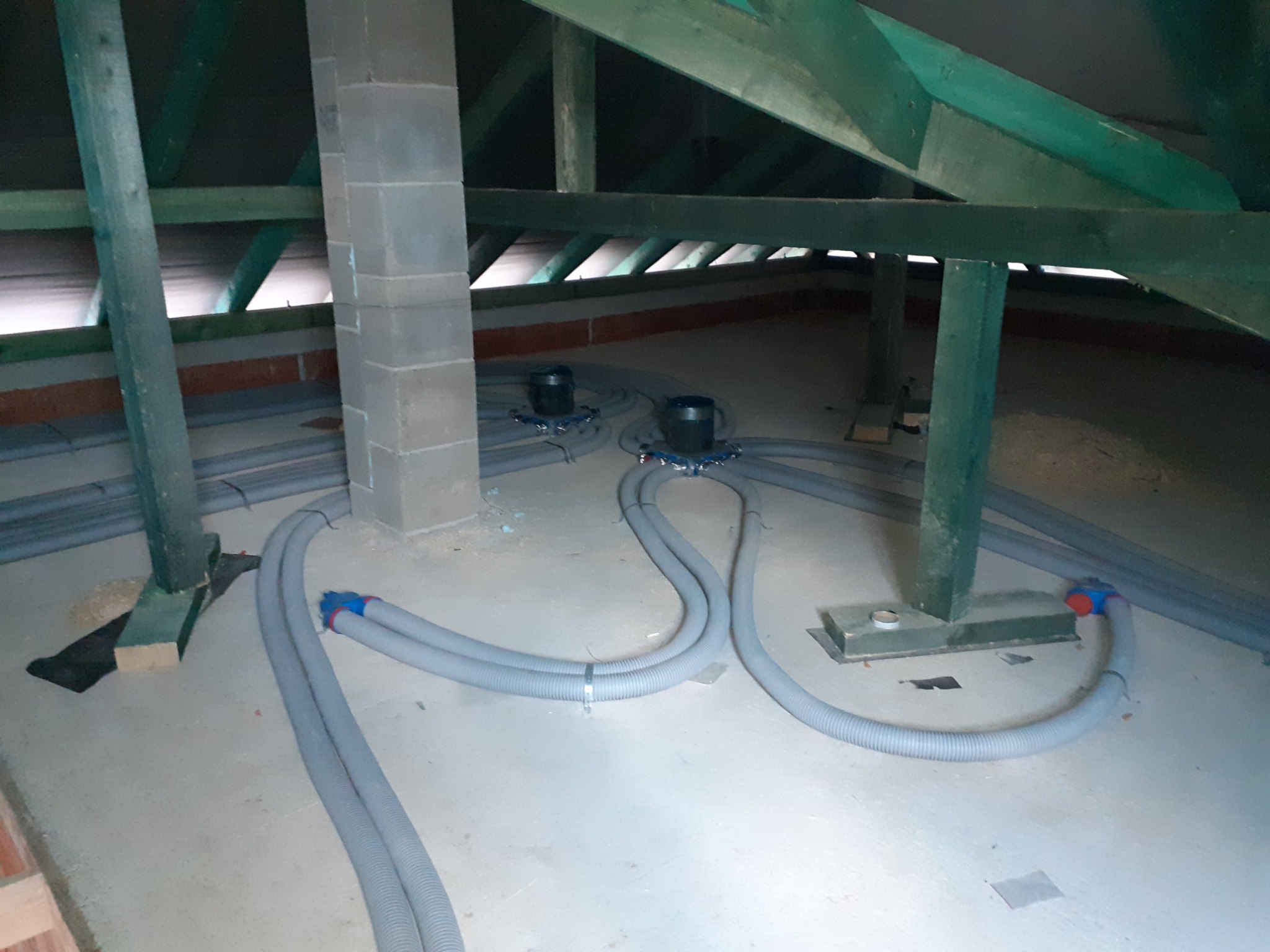 Szare, karbowane rury wentylacyjne prowadzone po podłodze strychu w kierunku dwóch pionowych kominów wentylacyjnych, widoczne elementy drewnianej konstrukcji dachu.