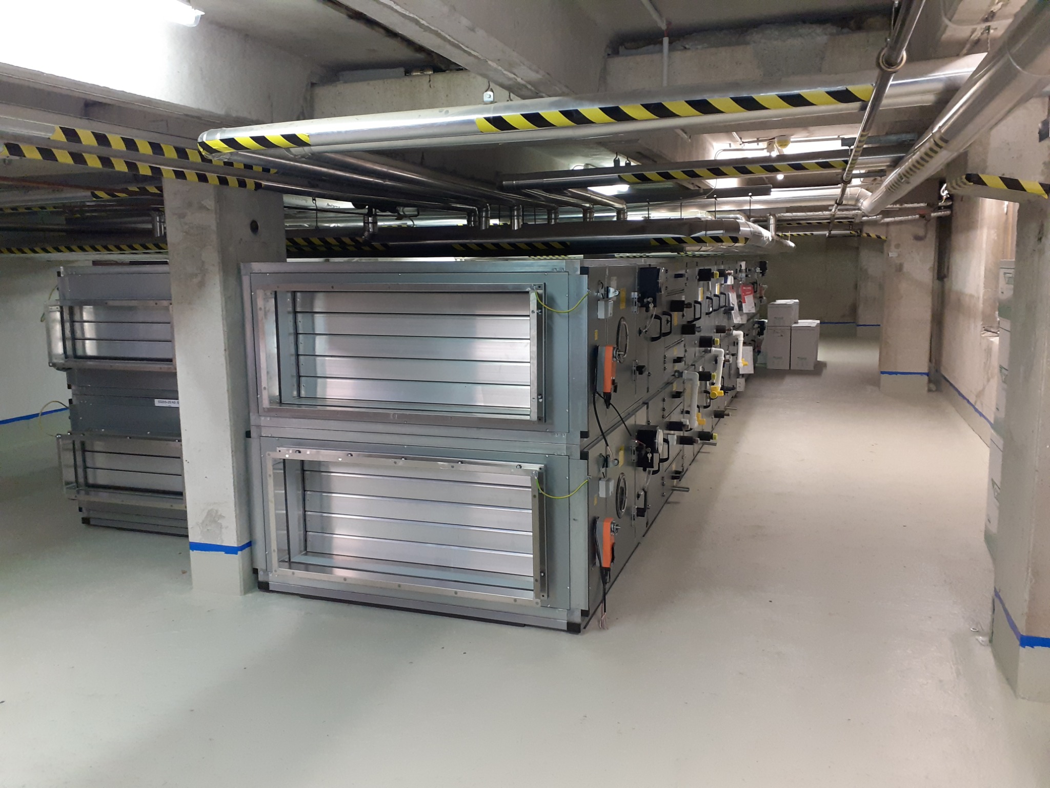 Długi rząd szarych, metalowych urządzeń wentylacyjnych z panelami i przyłączami, ustawionych w rzędzie w pomieszczeniu technicznym z betonowymi kolumnami i rurami pod sufitem oznaczonymi taśmą...