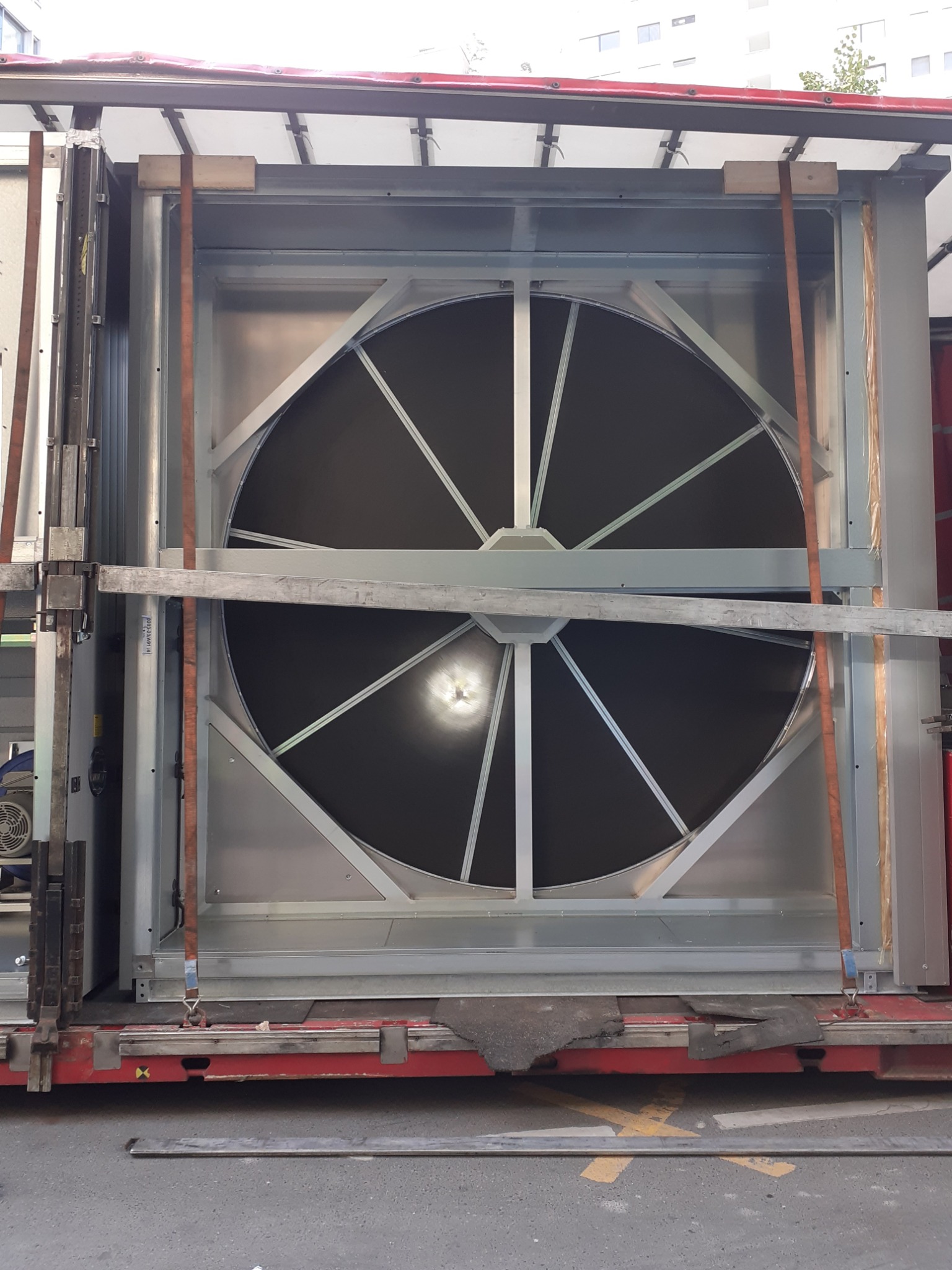 Duży, okrągły wentylator przemysłowy z metalową obudową, zabezpieczony pasami transportowymi, prawdopodobnie podczas transportu lub montażu.