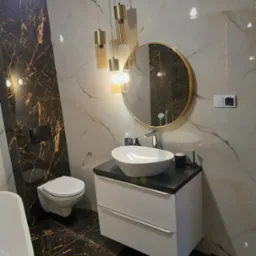 Nowoczesna łazienka z marmurowymi płytkami w odcieniach bieli i brązu, wisząca umywalka z szafką, okrągłe lustro w złotej ramie i designerskie lampy wiszące.