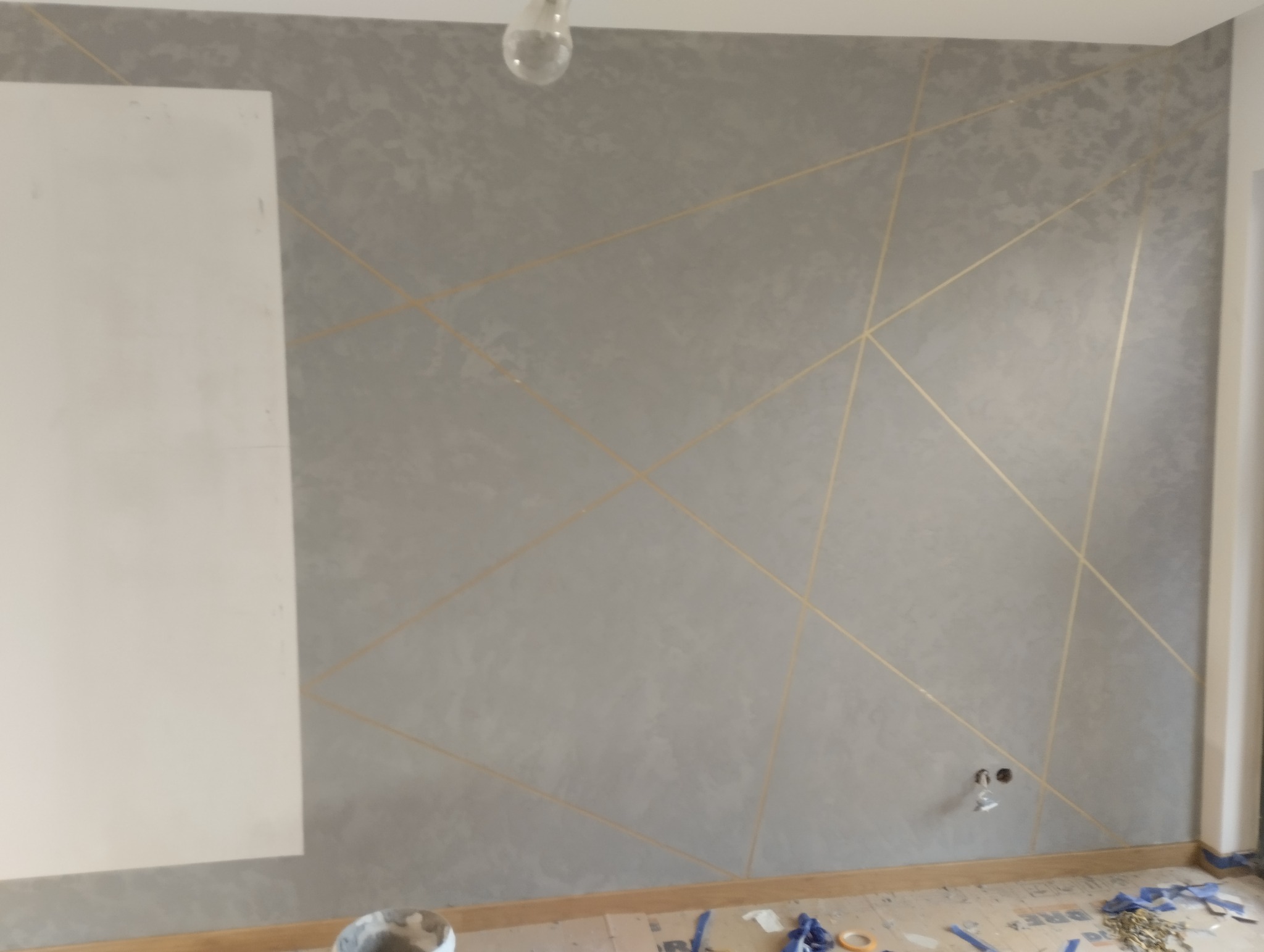 Ściana pokryta szarą gładzią z geometrycznym wzorem z cienkich, złotych linii; widoczny fragment niezabezpieczonej powierzchni oraz akcesoria malarskie na podłodze.