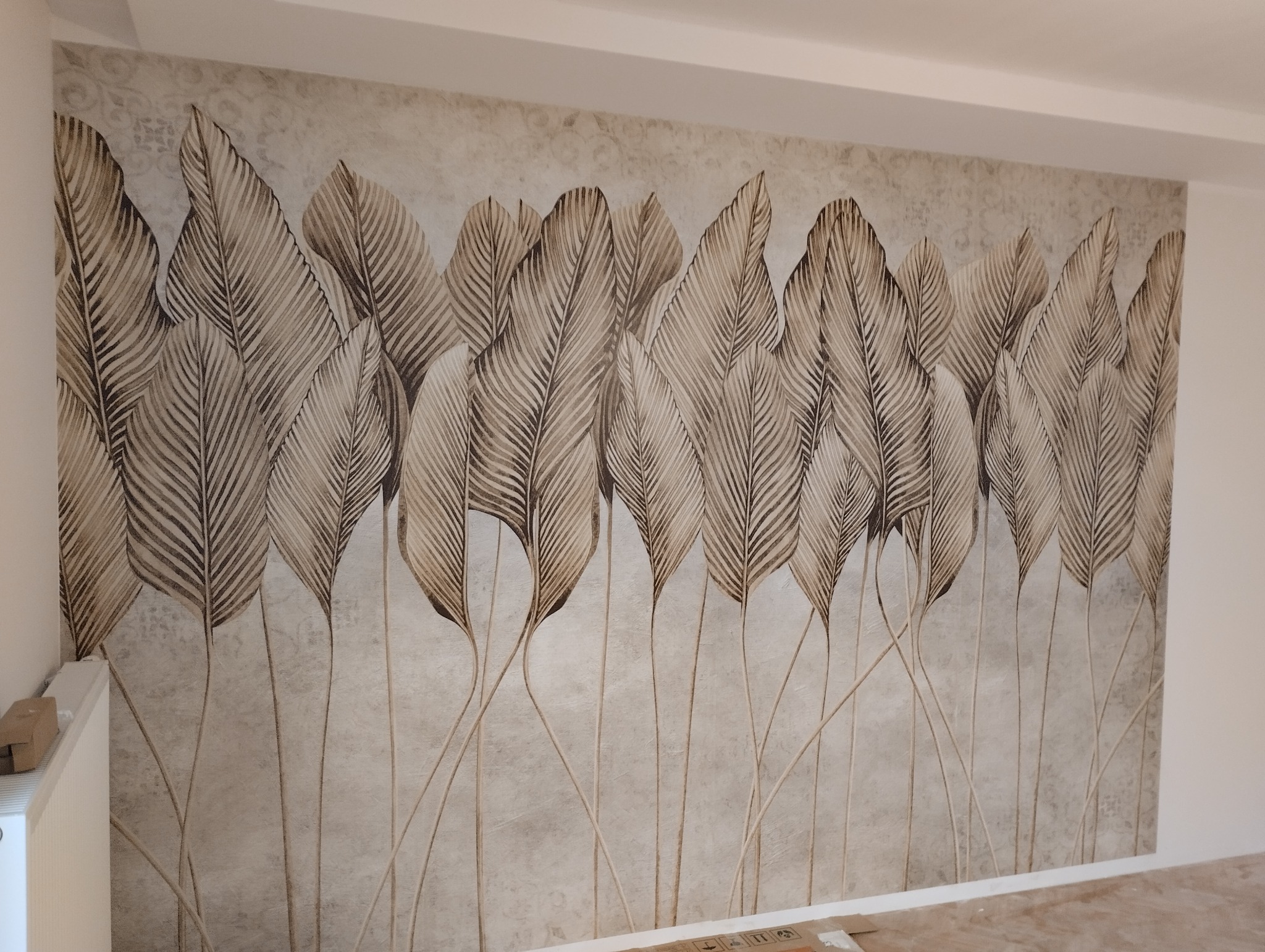 Ściana z tapetą w odcieniach beżu i brązu przedstawiającą motyw stylizowanych liści z wyraźną fakturą tła, widoczne fragmenty grzejnika i podłogi w trakcie prac wykończeniowych.
