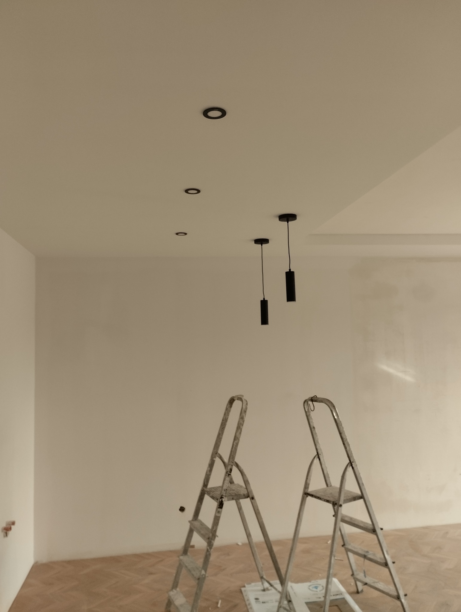 Montaż czarnych, cylindrycznych lamp wiszących i halogenowych w suficie podwieszanym; dwie aluminiowe drabiny rozstawione na parkiecie w trakcie prac wykończeniowych wnętrza.