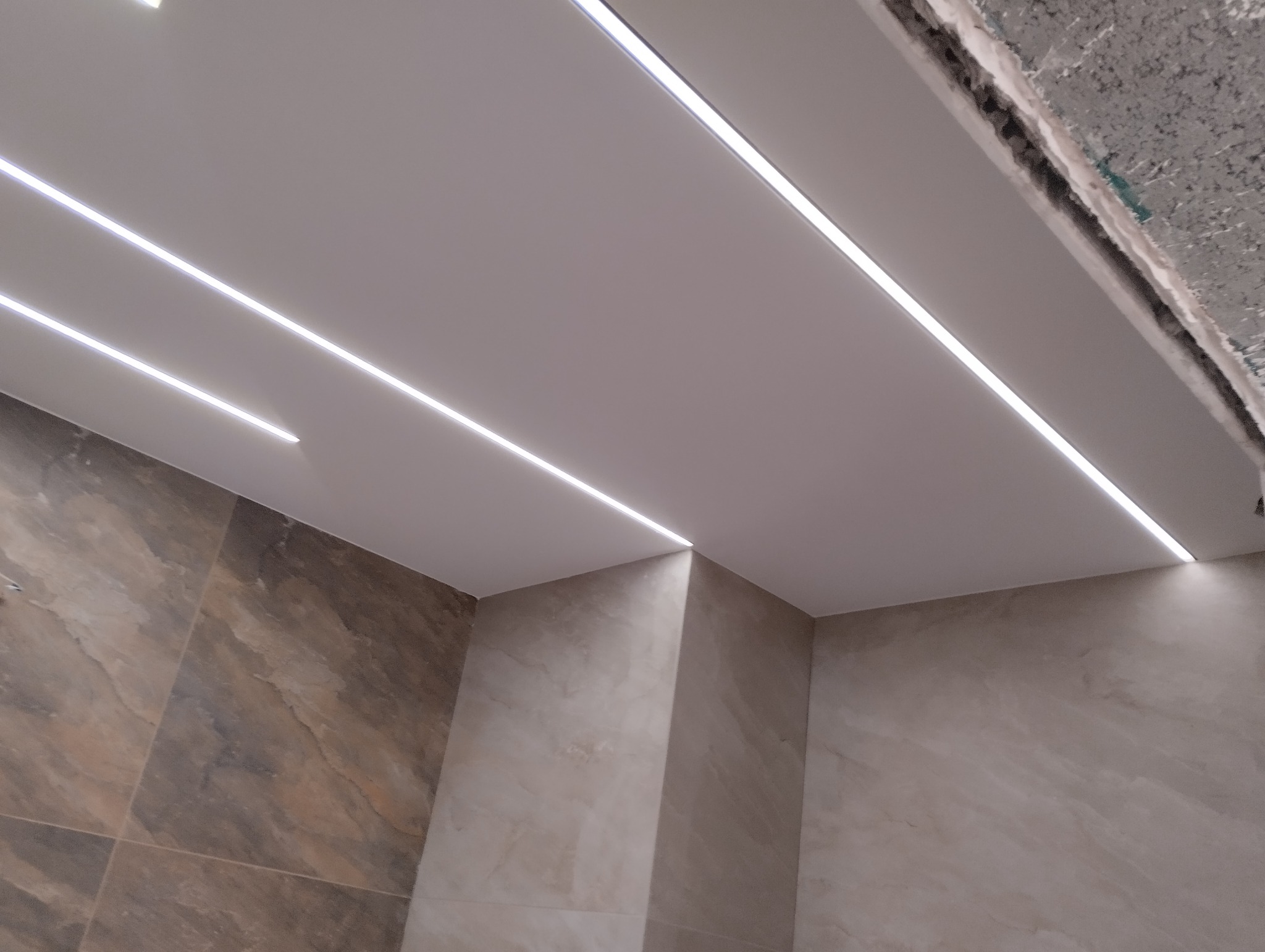 Nowoczesna łazienka w trakcie wykańczania z podwieszanym sufitem i liniowym oświetleniem LED, widoczne płytki imitujące marmur na ścianach, surowy betonowy strop w górnym rogu.