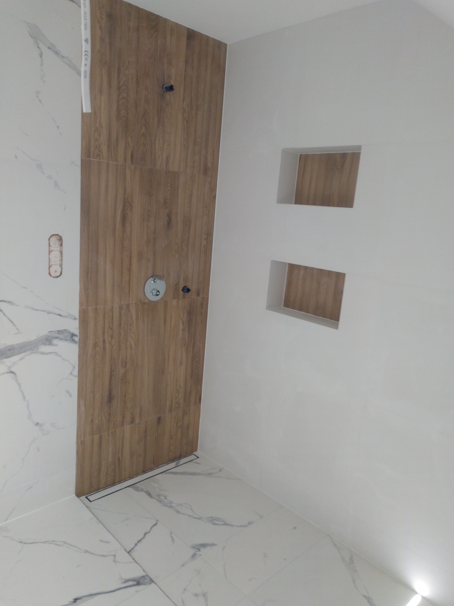 Wykończona kabina prysznicowa z drewnopodobnymi płytkami, odpływem liniowym i dwoma wnękami w ścianie, wykończona białymi płytkami imitującymi marmur.