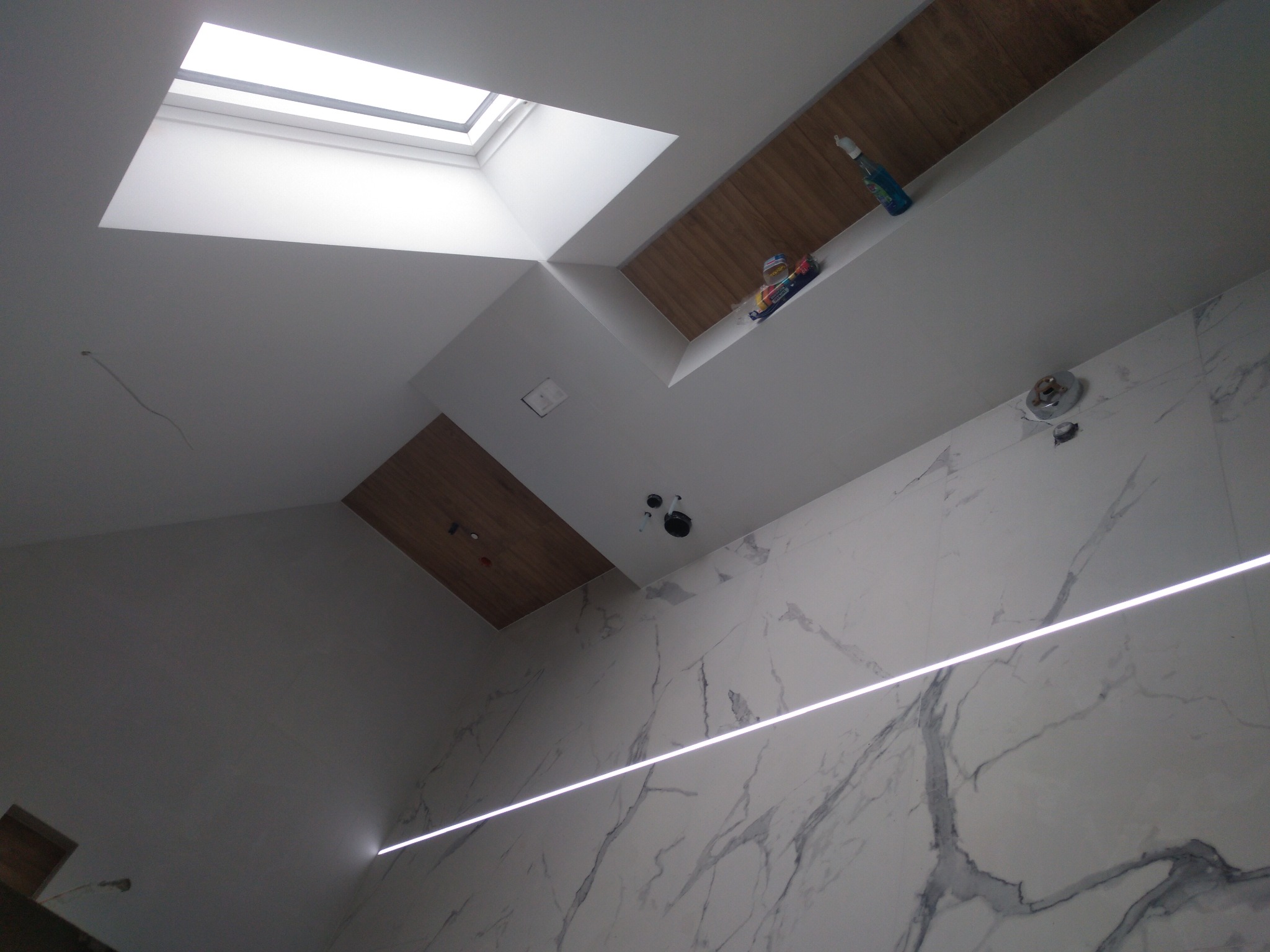 Skośny kadr na sufit i fragment ściany łazienki z marmurowymi płytkami, widoczne okno dachowe, oświetlenie LED i półka z zabawkami oraz środkiem czystości.