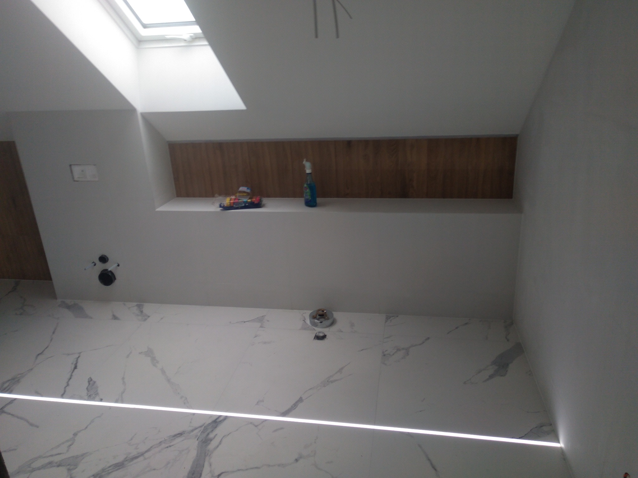 Nowoczesna łazienka z marmurowymi płytkami podłogowymi, oświetleniem LED w podłodze, drewnianą wnęką w ścianie i oknem dachowym, z widocznymi elementami instalacji wodnej.