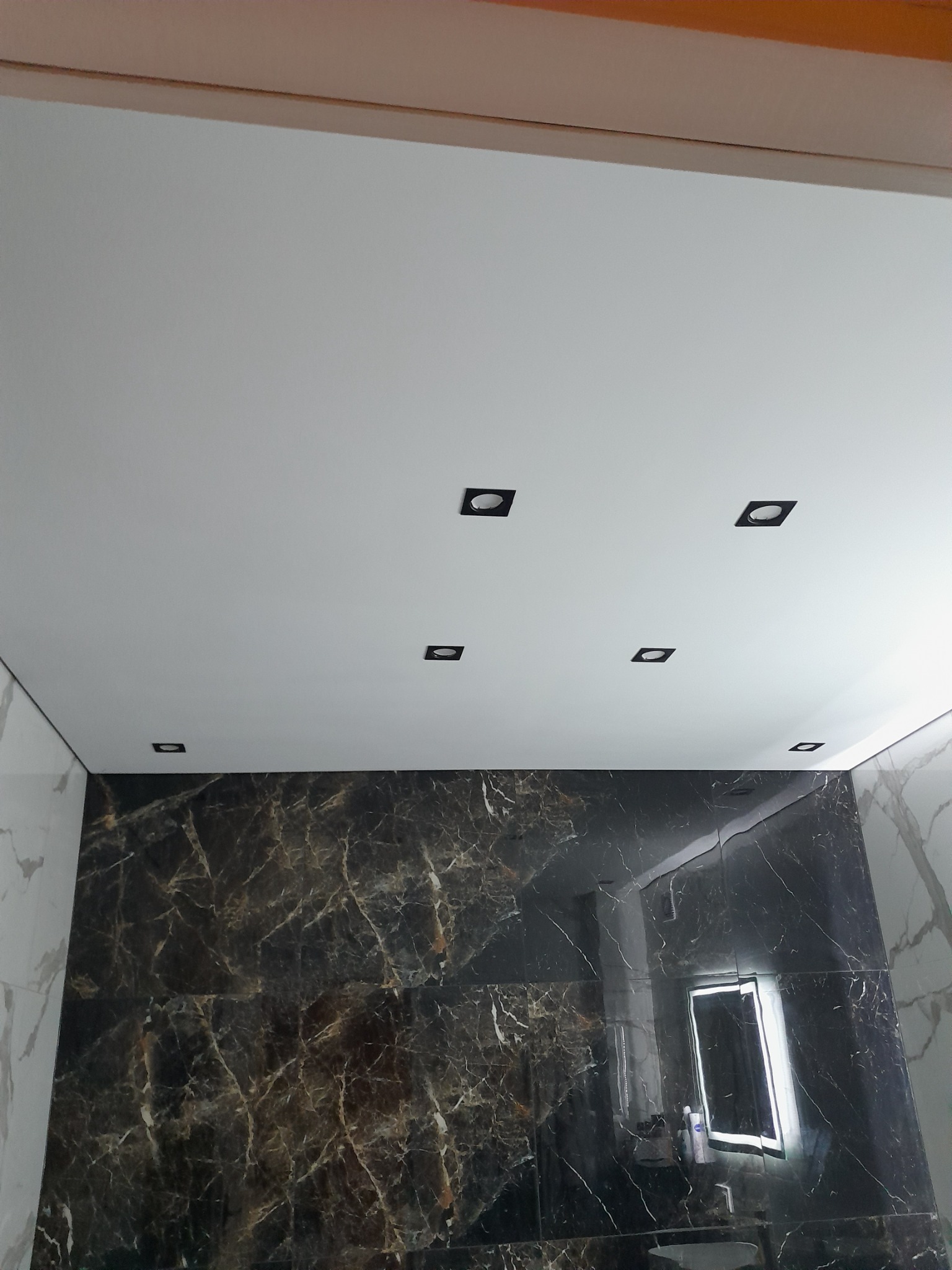 Częściowe ujęcie wykończonej łazienki z czarnymi płytkami imitującymi marmur, białym sufitem podwieszanym z sześcioma czarnymi, kwadratowymi lampami punktowymi i podświetlanym lustrem.