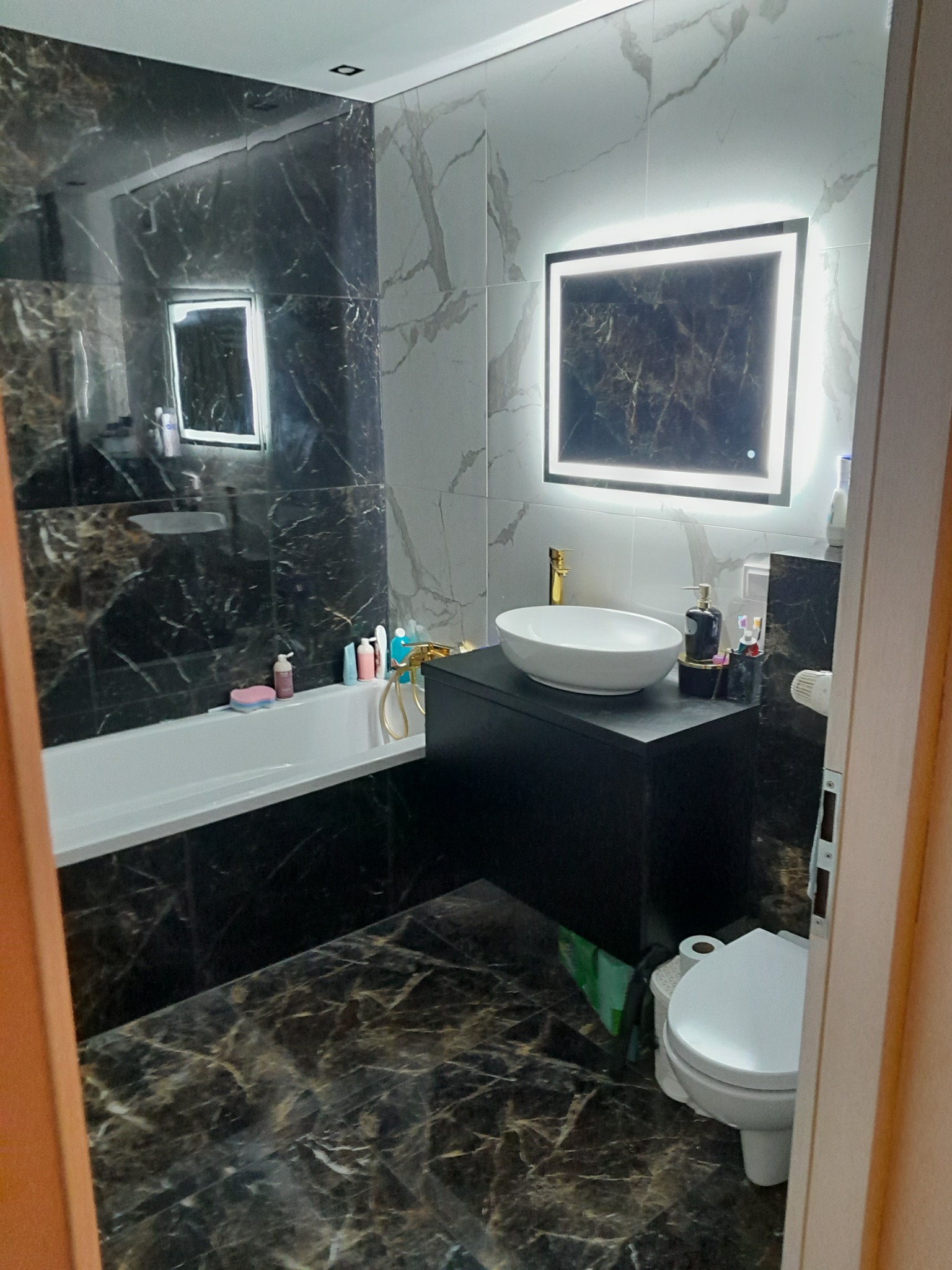 Nowoczesna łazienka z czarno-białymi marmurkowymi płytkami, podświetlanym lustrem, białą umywalką na czarnej szafce i białą wanną.