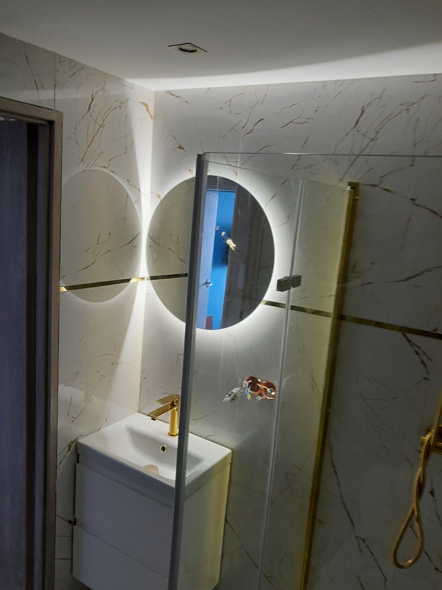 Wykończona łazienka z marmurowymi płytkami, okrągłym lustrem z podświetleniem LED, białą szafką pod umywalką i złotą armaturą, widoczne przyłącza elektryczne.