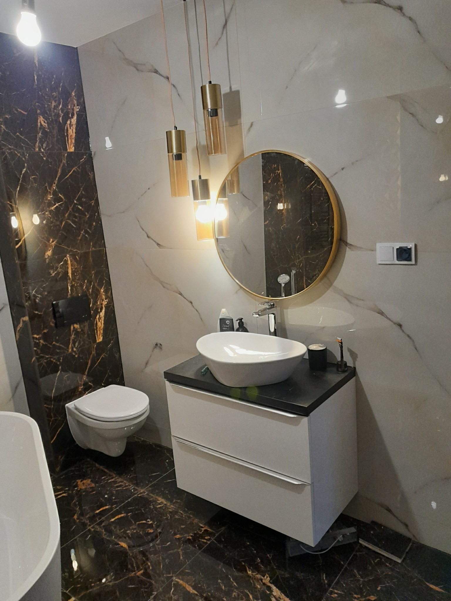 Nowoczesna łazienka z marmurowymi płytkami w odcieniach bieli i brązu, wisząca umywalka z szafką, okrągłe lustro w złotej ramie i designerskie lampy wiszące.
