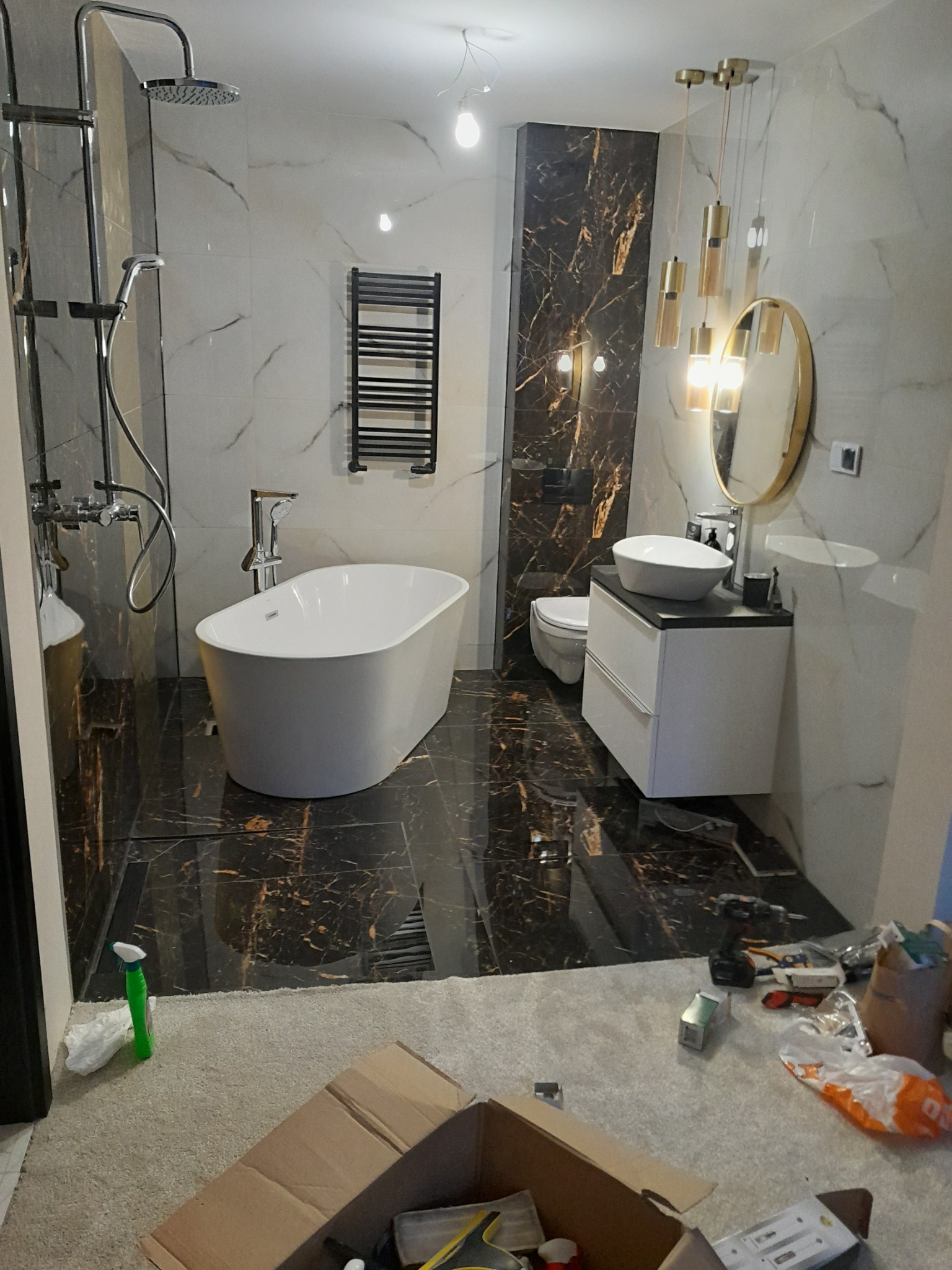 Nowoczesna łazienka w Kurzętniku z marmurowymi płytkami, wolnostojącą wanną i złotymi akcentami oświetleniowymi, w trakcie prac wykończeniowych, widoczne narzędzia i kartony na podłodze.