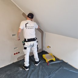 BUD-MAT - Pracownik w białych spodniach i koszulce z logo firmy maluje ścianę w skośnym pomieszczeniu, widoczny wałek i kuweta z farbą, podłoga zabezpieczona folią.