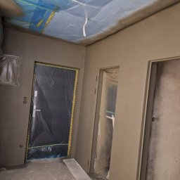 BUD-MAT - Przygotowanie do malowania ścian w pomieszczeniu: ściany pokryte świeżą gładzią, drzwi zabezpieczone folią malarską, sufit osłonięty niebieską folią ochronną.