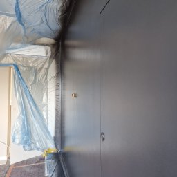 BUD-MAT - Świeżo pomalowana, ciemnoszara ściana z widocznymi zabezpieczeniami folią malarską na klimatyzatorze i framudze drzwi, widoczne gniazdko elektryczne, wykonane z bliskiej odległości.