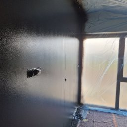 BUD-MAT - Świeżo pomalowana na ciemny kolor ściana z widocznym gniazdkiem elektrycznym, zabezpieczona folią okienna i fragment podłogi.