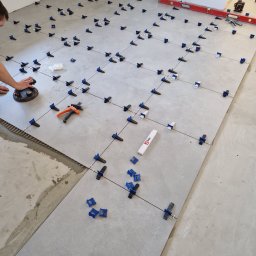 BUD-MAT - Układanie szarych płytek podłogowych z użyciem systemu poziomowania, widoczne przyssawka do przenoszenia płytek, poziomica i miarka składana.